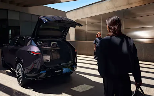 Mazda CX-6e elektromos SUV nyitott csomagtartóval modern épület előtt, utazótáskákkal a csomagtérben