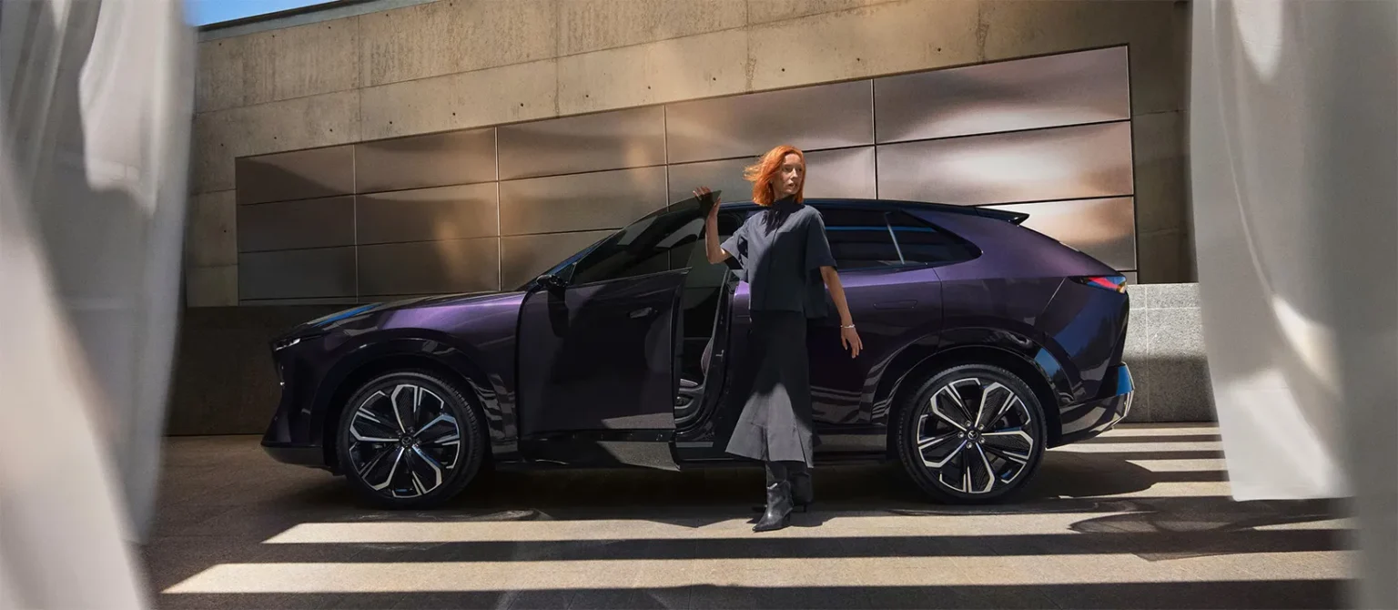 Mazda CX-6e elektromos SUV oldalnézetből, egy nő áll a nyitott ajtó mellett modern épület előtt