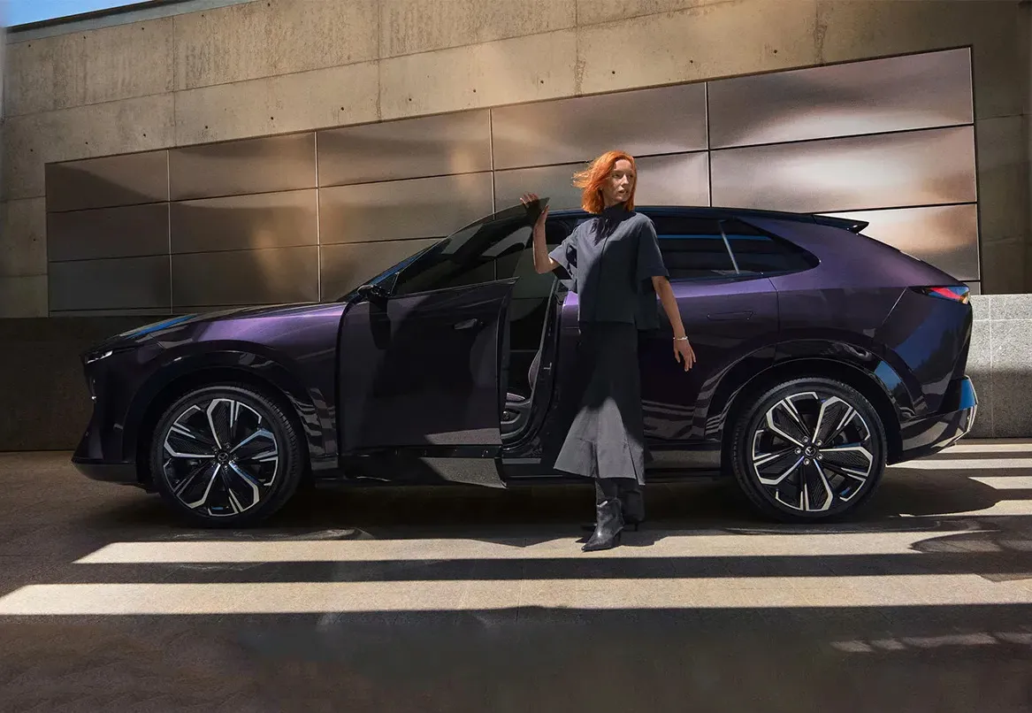 Mazda CX-6e elektromos SUV oldalnézetből, egy nő áll a nyitott ajtó mellett modern épület előtt